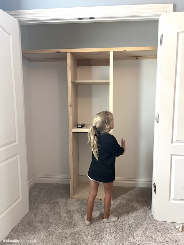 Process shot of girls closet. Wooden frame with top shelf. Little girl looking around it.