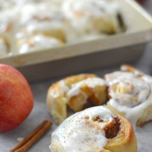 Profile image of Apple Cinnamon Rolls. One cinnamon roll close up, remaining rolls in pan behind.